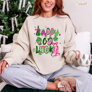 A woman wearing a white sweatshirt with a colorful graphic design and text, sitting on a couch and holding a mug.