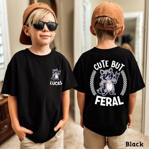 A young boy wearing a black t-shirt with a cartoon raccoon and the text "CUTE BUT FERAL" printed on it.