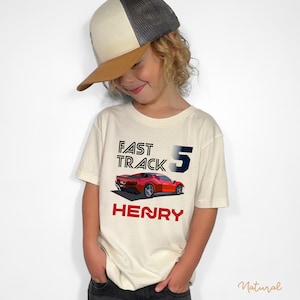 May include: A cream-colored t-shirt with the words "FAST TRACK 5" and the name "HENRY" in red. A red sports car graphic is below the text. The child is wearing a trucker hat with a brown brim and a gray mesh back.