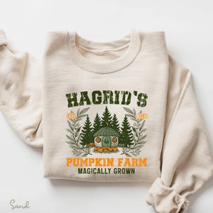 May include: Cream-colored sweatshirt featuring a green graphic with the text "Hagrid's Pumpkin Farm" and the phrase "Magically Grown." The design includes a small house, pumpkins, and trees. The sweatshirt has a crew neck and long sleeves.