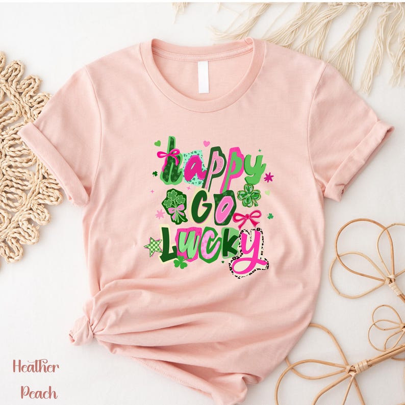 A pink t-shirt with a colorful graphic design featuring the text "Happy Go Lucky" surrounded by various Irish symbols and elements.