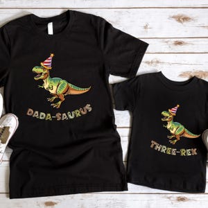 two black t-shirts with a dinosaur wearing a party hat and the text "Dada-Saurus" printed on them.