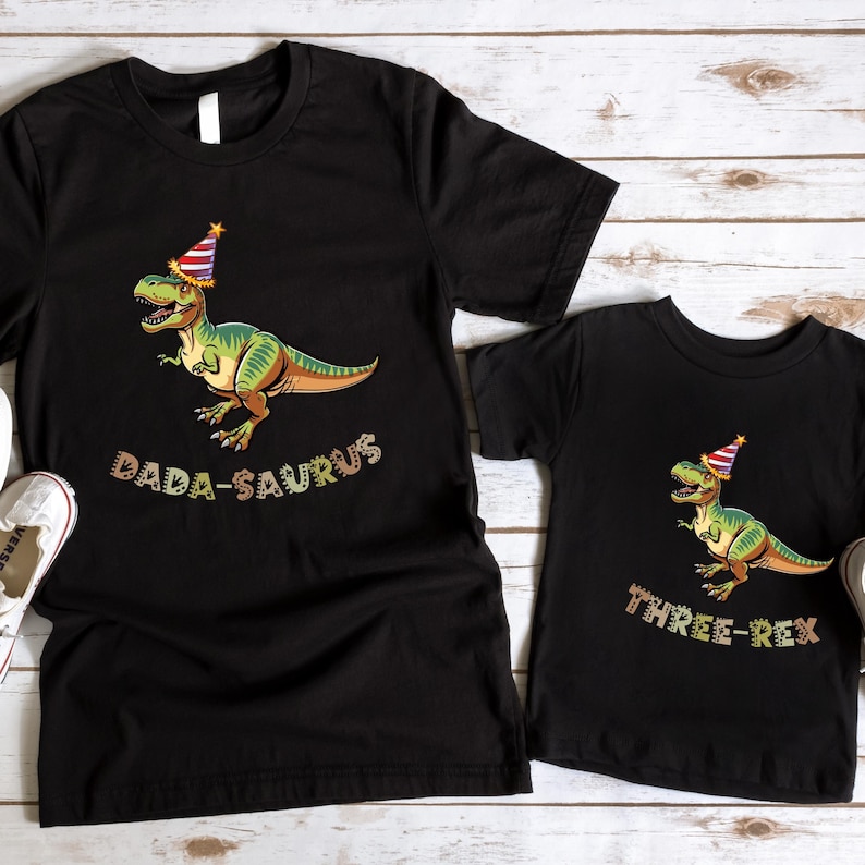 two black t-shirts with a dinosaur wearing a party hat and the text "Dada-Saurus" printed on them.