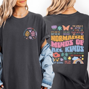 May include: Dark gray t-shirt with the text "Normalize Minds of All Kinds" and colorful floral graphics on the front and back. The back also features additional text and graphics, including a rainbow and butterfly.