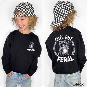 A young boy wearing a black sweatshirt with a cartoon raccoon and the text "CUTE BUT FERAL" printed on it.