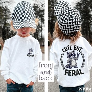 A young child wearing a white shirt with a checkered hat and a graphic of a raccoon, along with the text "CUTE BUT FERAL" printed on the back.