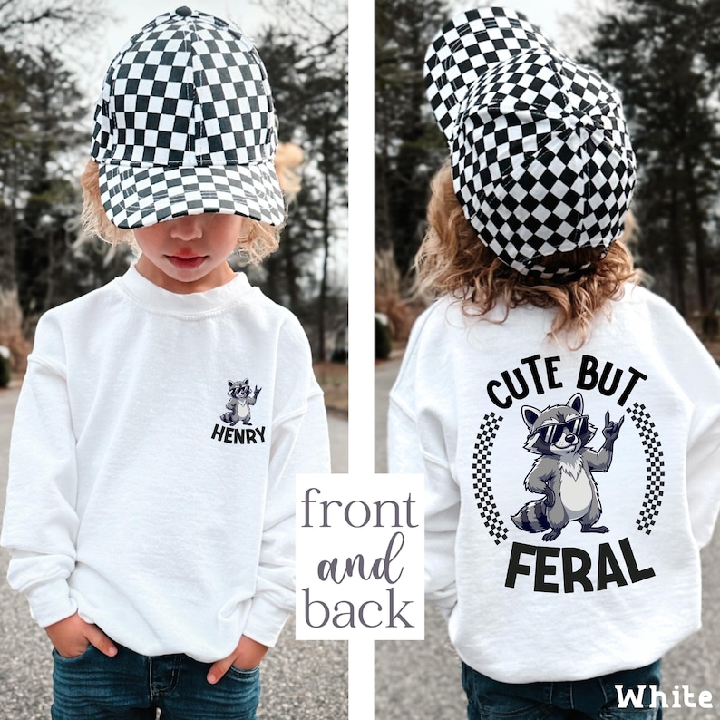 A young child wearing a white shirt with a checkered hat and a graphic of a raccoon, along with the text "CUTE BUT FERAL" printed on the back.