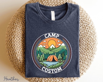 May include: A heather navy blue t-shirt featuring a circular graphic with the text CAMP COSTOM. The design depicts a landscape with a tent, a lake, green pine trees, snow-capped mountains, and a yellow sun under a sky with clouds.