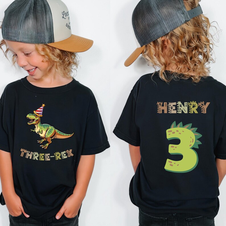 A young boy wearing a black t-shirt with a dinosaur graphic and the text "THREE REX" and the number "3" on the back, along with a baseball cap.