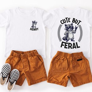 a pair of children's clothing items, including a white t-shirt with a raccoon graphic and the phrase "CUTE BUT FERAL", a pair of brown shorts, and a pair of checkered shoes.