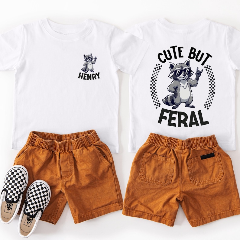 a pair of children's clothing items, including a white t-shirt with a raccoon graphic and the phrase "CUTE BUT FERAL", a pair of brown shorts, and a pair of checkered shoes.
