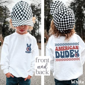 A young child wearing a white sweatshirt with a checkered hat and a peace sign on the front, and a white sweatshirt with the text "American Dude" on the back.