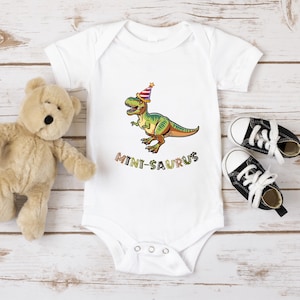 A white onesie with a cartoon dinosaur wearing a party hat, a teddy bear, and a pair of black and white sneakers are arranged on a wooden surface.