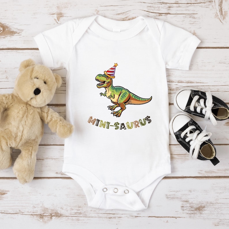 A white onesie with a cartoon dinosaur wearing a party hat, a teddy bear, and a pair of black and white sneakers are arranged on a wooden surface.