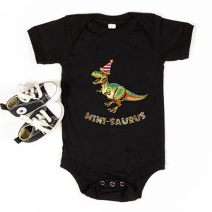 A black baby onesie with a cartoon dinosaur wearing a party hat and the text "MINI-SAURUS" printed on it, along with a pair of black and white sneakers.