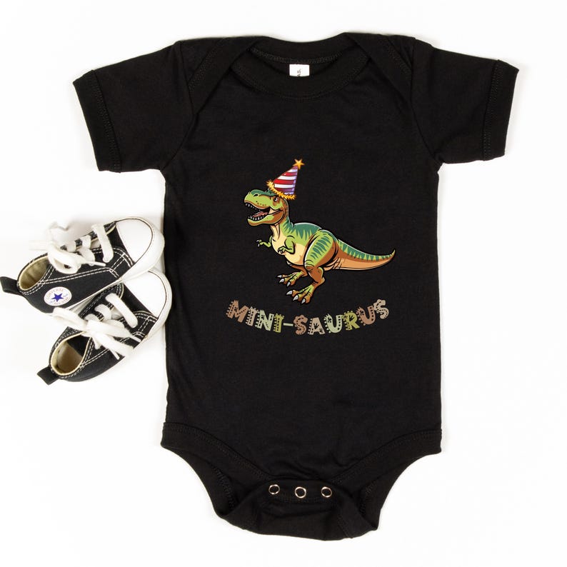 A black baby onesie with a cartoon dinosaur wearing a party hat and the text "MINI-SAURUS" printed on it, along with a pair of black and white sneakers.