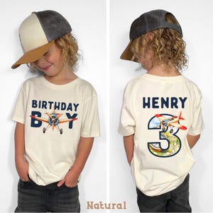 May include: Cream-colored t-shirt with a navy blue "BIRTHDAY BOY" graphic and an airplane illustration. The back of the shirt features the name "HENRY" and a number "3" with an airplane design. The shirt is worn by a child.