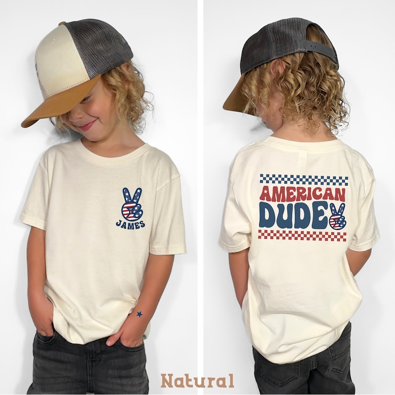 A young boy wearing a white t-shirt with a peace sign and the text "American Dude" printed on it, along with a baseball cap.