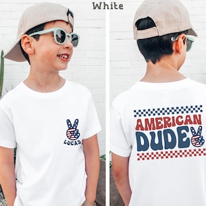 A young boy wearing a white t-shirt with a peace sign and the text "American Dude" printed on it, smiling and posing in front of a white brick wall.