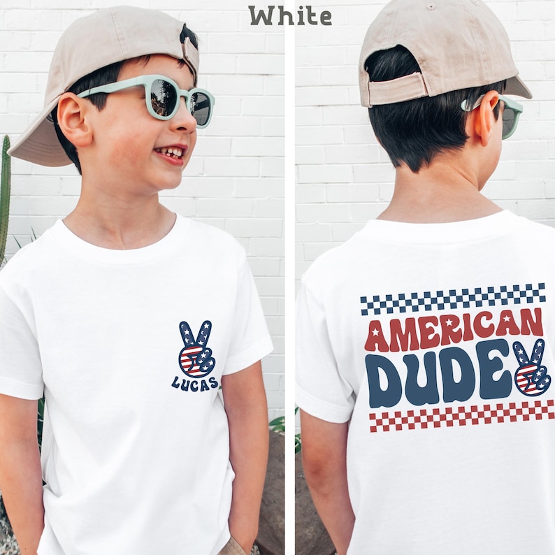 A young boy wearing a white t-shirt with a peace sign and the text "American Dude" printed on it, smiling and posing in front of a white brick wall.