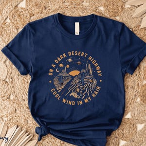 Hand-drawn graphic t-shirt featuring a dreamy desert landscape at sunset with surreal elements like planets in the sky, cacti, and animal skulls. The design includes the iconic lyric “On a Dark Desert Highway, Cool Wind in My Hair” from the classic Eagles song Hotel California. A perfect blend of retro, vintage, and boho aesthetics for music lovers and free spirits.