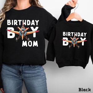 A woman wearing a black sweatshirt with a graphic of an airplane and the text "BIRTHDAY BOY MOM" printed on it, and a smaller black sweatshirt with the same text and graphic.