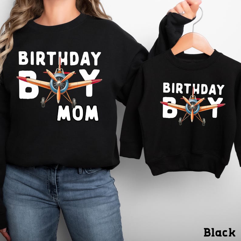 A woman wearing a black sweatshirt with a graphic of an airplane and the text "BIRTHDAY BOY MOM" printed on it, and a smaller black sweatshirt with the same text and graphic.
