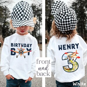 A young child wearing a white sweatshirt with a checkered hat and a white shirt with a plane design, standing in front of a background of trees.