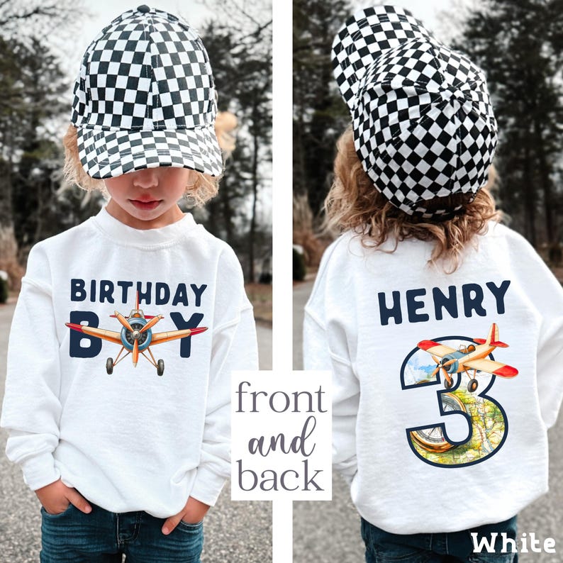 A young child wearing a white sweatshirt with a checkered hat and a white shirt with a plane design, standing in front of a background of trees.