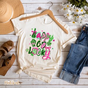 A casual outfit consisting of a white t-shirt with a colorful graphic, blue jeans, and brown sandals is laid out on a wooden surface, along with a straw hat and a bouquet of white flowers.