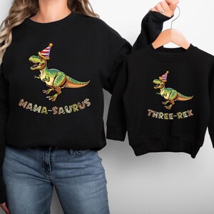 A woman wearing a black sweatshirt with a graphic of a dinosaur wearing a party hat and the text "MAMA-SAURUS" printed on it, and a smaller version of the same sweatshirt hanging on a hanger.