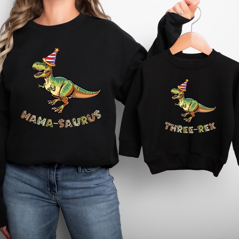 A woman wearing a black sweatshirt with a graphic of a dinosaur wearing a party hat and the text "MAMA-SAURUS" printed on it, and a smaller version of the same sweatshirt hanging on a hanger.