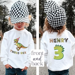 A young child wearing a white sweatshirt with a dinosaur and a checkered hat, and a white sweatshirt with the name "HENRY" and the number "3" printed on it.