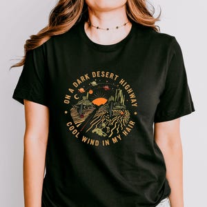 Hand-drawn graphic t-shirt featuring a dreamy desert landscape at sunset with surreal elements like planets in the sky, cacti, and animal skulls. The design includes the iconic lyric “On a Dark Desert Highway, Cool Wind in My Hair” from the classic Eagles song Hotel California. A perfect blend of retro, vintage, and boho aesthetics for music lovers and free spirits.