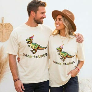 May include: Two cream-colored t-shirts with matching dinosaur graphics. One shirt reads "DADA-SAURUS" and the other reads "MAMA-SAURUS". The dinosaur is green, orange, and yellow, wearing a party hat. The shirts are short-sleeved.