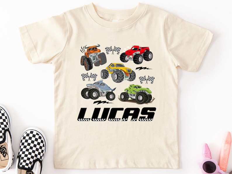 A white t-shirt with a graphic design of various monster trucks and checkered flags, along with the name "LUCAS" printed below.