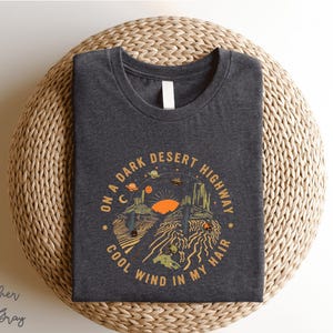 Hand-drawn graphic t-shirt featuring a dreamy desert landscape at sunset with surreal elements like planets in the sky, cacti, and animal skulls. The design includes the iconic lyric “On a Dark Desert Highway, Cool Wind in My Hair” from the classic Eagles song Hotel California. A perfect blend of retro, vintage, and boho aesthetics for music lovers and free spirits.