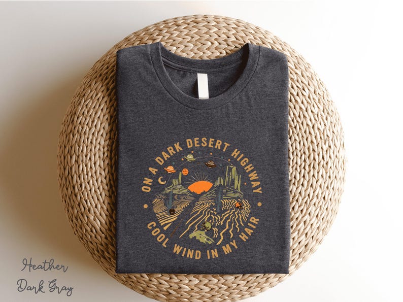 Hand-drawn graphic t-shirt featuring a dreamy desert landscape at sunset with surreal elements like planets in the sky, cacti, and animal skulls. The design includes the iconic lyric “On a Dark Desert Highway, Cool Wind in My Hair” from the classic Eagles song Hotel California. A perfect blend of retro, vintage, and boho aesthetics for music lovers and free spirits.