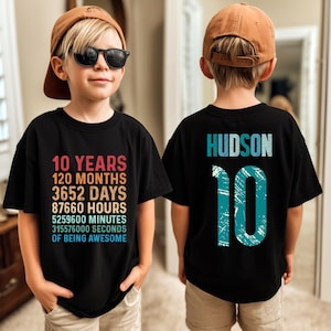 May include: Black t-shirt with the text "10 YEARS, 120 MONTHS, 3652 DAYS, 87660 HOURS, 5259600 MINUTES, 315576000 SECONDS OF BEING AWESOME" in a retro color scheme. The back of the shirt has the name "HUDSON" and the number "10".