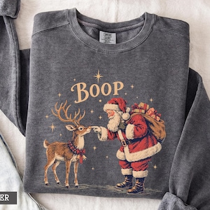 Puede incluir: Una sudadera de cuello redondo gris carbón con un gráfico de Santa Claus tocando la nariz de un reno. El texto BOOP está impreso encima en una fuente vintage, rodeado de pequeñas estrellas. La sudadera tiene cuello y puños acanalados.