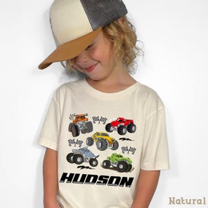 A young child wearing a baseball cap and a t-shirt with a graphic of colorful monster trucks.