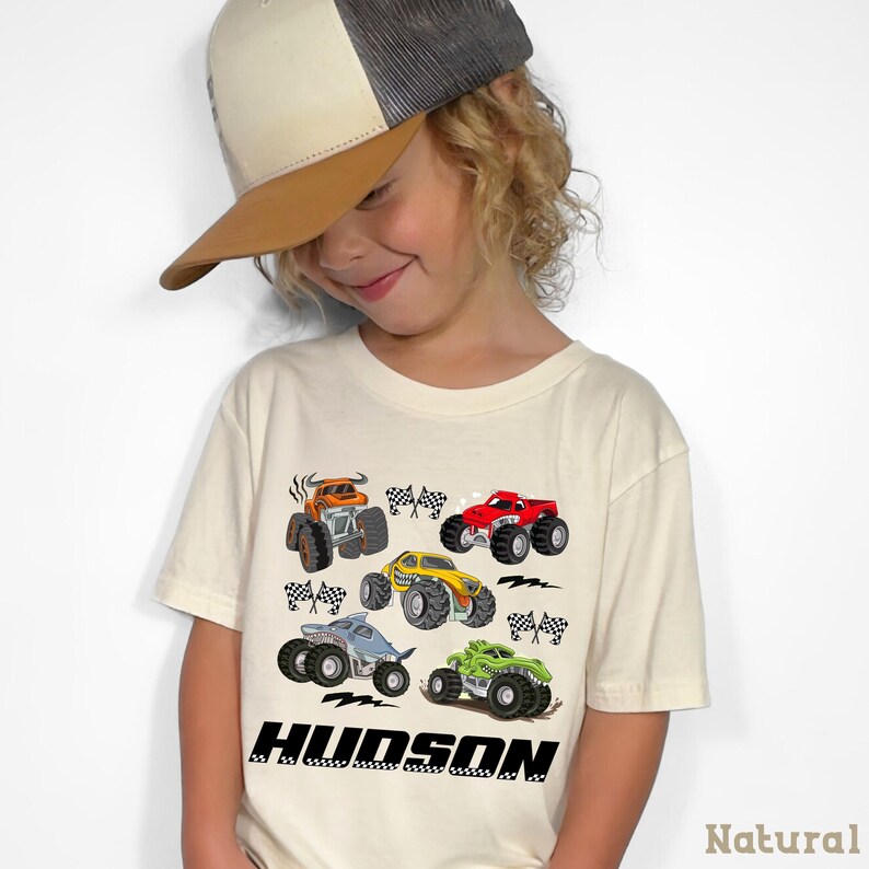 A young child wearing a baseball cap and a t-shirt with a graphic of colorful monster trucks.
