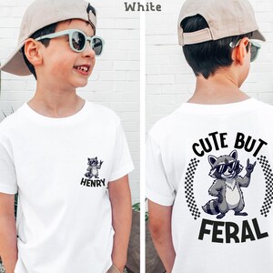 A young boy wearing a white t-shirt with a graphic of a raccoon and the text "CUTE BUT FERAL" printed on it.
