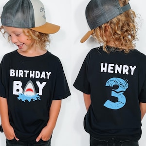 May include: Black t-shirt with the words "BIRTHDAY BAY" and a shark graphic. The back of the shirt has the name "HENRY" and the number "3" with a wave design. The child is wearing a trucker hat.