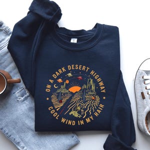 Hand-drawn graphic t-shirt featuring a dreamy desert landscape at sunset with surreal elements like planets in the sky, cacti, and animal skulls. The design includes the iconic lyric “On a Dark Desert Highway, Cool Wind in My Hair” from the classic Eagles song Hotel California. A perfect blend of retro, vintage, and boho aesthetics for music lovers and free spirits.