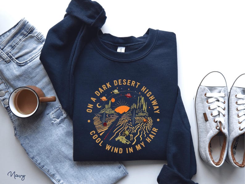 Hand-drawn graphic t-shirt featuring a dreamy desert landscape at sunset with surreal elements like planets in the sky, cacti, and animal skulls. The design includes the iconic lyric “On a Dark Desert Highway, Cool Wind in My Hair” from the classic Eagles song Hotel California. A perfect blend of retro, vintage, and boho aesthetics for music lovers and free spirits.