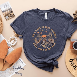 Hand-drawn graphic t-shirt featuring a dreamy desert landscape at sunset with surreal elements like planets in the sky, cacti, and animal skulls. The design includes the iconic lyric “On a Dark Desert Highway, Cool Wind in My Hair” from the classic Eagles song Hotel California. A perfect blend of retro, vintage, and boho aesthetics for music lovers and free spirits.