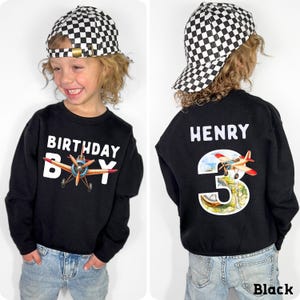 A young boy wearing a black sweatshirt with a checkered hat and a birthday shirt with a plane design, smiling at the camera.