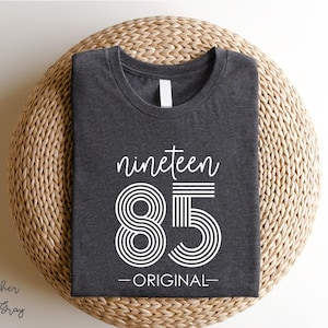 May include: Dark gray t-shirt with the text "nineteen 85 - ORIGINAL" in white. The number 85 is designed with concentric circles and horizontal lines. The shirt is folded neatly on a woven surface.