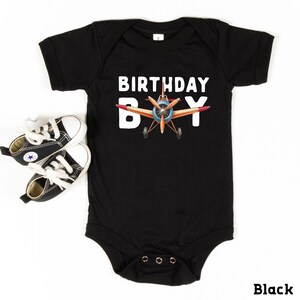 A black baby onesie with the text "BIRTHDAY BOY" printed on it, along with an illustration of an airplane. The onesie is placed next to a pair of black and white sneakers.